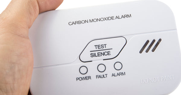 A close-up of someone holding a white carbon monoxide alarm. The alarm has test, silence, power, fault, and alarm buttons.