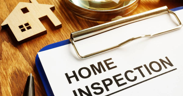 A clipboard with a paper that reads "Home Inspection" sits on a desk near a small wooden house and a magnifying glass.