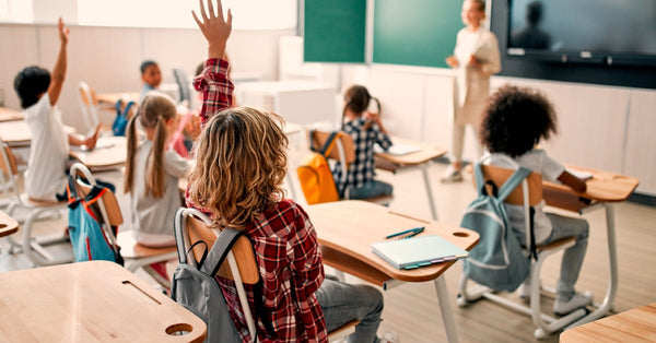 Young children sit at desks in a classroom as they face the teacher. Two of the kids raise their hands.