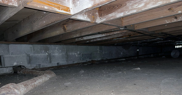 A large metal pipe rests on the dirt floor of a home's crawl space and exits the cinderblock foundation wall.