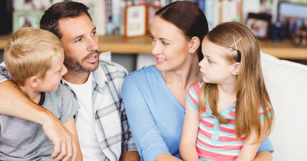 A family of four sit together on the sofa at home. The dad explains something as the mom, daughter, and son listen.