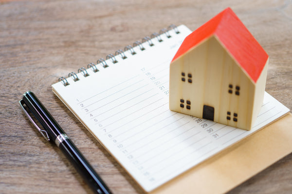 A wooden model of a house with a red roof sits on top of a spiral notebook opened to a blank page. A pen sits nearby.