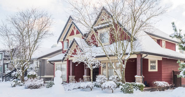 A snowman sits in the front yard of a red craftsman-style house. The landscaping and sidewalk are covered in snow.