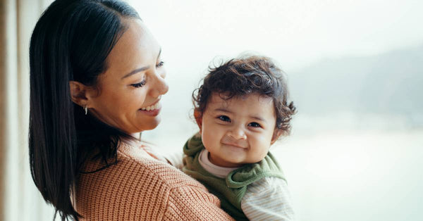 A smiling mother looks down the baby in her arms while standing near a picture window. The baby grins at the camera.